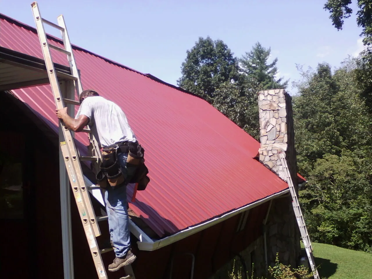 Roofer on a ladder beside a red metal roof and stone chimney — Soffit & Fascia Repair by Roofing Brothers in Cummings